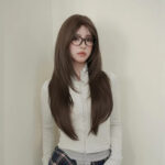 Young woman with long dark brown hair and glasses posing against a pale wall, wearing a light gray zip-up hoodie.