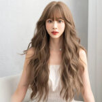Young woman with long, wavy light brown hair and bangs, wearing a white strapless top indoors against a light wall.