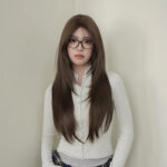 Woman with long brown hair and glasses stands against a beige wall in a light gray zip-up sweater and plaid skirt.