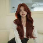 Young woman with long, wavy red hair wearing a white blouse, sitting indoors and looking at the camera.