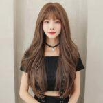 Portrait of a young woman with long wavy brown hair and bangs, wearing a black cropped top and a star pendant choker in front of a light background.