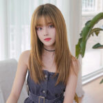 Portrait of a young woman with straight light-brown hair wearing a denim dress and a black choker, indoors near a plant.