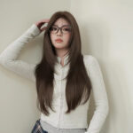 Woman with long brown hair and glasses wearing a light gray zip-up sweater, posing against a pale wall.