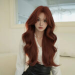 Portrait of a woman with long wavy red hair wearing a white blouse and pearl necklace, seated in a bright room.