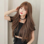 Young woman with long wavy brown hair wearing a black crop top and choker, posing beside a light wall.