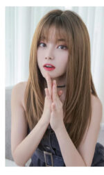 Young woman with long light-brown hair and bangs, hands pressed together near her chest, looking softly at the camera in a bright room.