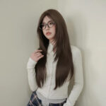 Woman with long brown hair and glasses wearing a light knit top, posing against a pale wall with hand in hair.