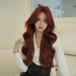 Woman with long, wavy red hair wearing a white blouse and pearl necklace, sitting on a light-colored couch indoors.