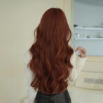 Back view of a person with long, wavy auburn hair cascading down to the mid back, wearing a white top indoors.