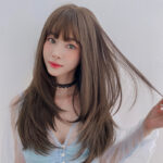 Portrait of a young woman with long brown hair, blue top, and a black lace choker, looking at the camera against a pale background.