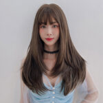 Portrait of a young woman with long brown hair and bangs, wearing a black lace choker and a light blue sheer-top, posing against a pale wall.