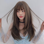 Portrait of a young woman with long brown hair and bangs, wearing a light blue top with sheer sleeves, looking to the side while pulling her hair.