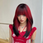Young woman with red and black layered hair and bangs, wearing a red sleeveless top and silver necklace, posing indoors by a light wall.