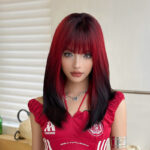 Young woman with red-to-black ombré hair and straight bangs, wearing a red off-shoulder top and layered necklace, looking at the camera.