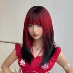 Portrait of a young woman with red and black straight hair, wearing a red athletic top with logos and a silver butterfly necklace, posing with hands on her hips.