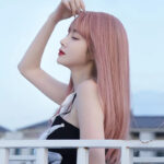 Side profile of a woman with long pastel pink hair posing with her eyes closed, one hand touching her hair against a pale blue sky.