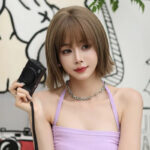 Portrait of a young woman with a short brown bob holding a black camera, wearing a lavender halter top and a silver chain necklace, indoors with abstract wall art in the background.