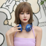 Portrait of a young woman with short light brown hair wearing blue over-ear headphones around her neck, in a lavender tank top, looking to the side against a graffiti background