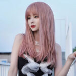 Young woman with long pink hair and red lipstick posing outdoors, wearing a black dress with a white sheer bow, looking to the left.