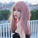 A young woman with long pastel pink hair poses on a balcony, looking toward the camera with a soft expression.