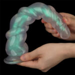 Glow-in-the-dark flexible silicone sex toy twisted into a spiral, held by two hands against a black background.