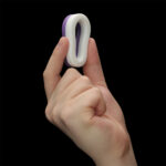 Close-up of a hand pinching a small white and purple loop-shaped silicone toy between the thumb and index finger against a black background.